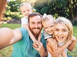 Nature, selfie and portrait of a happy family on a picnic together in outdoor green garden. Happy, smile and parents playing, hugging and bonding with children outside in backyard or park in canada